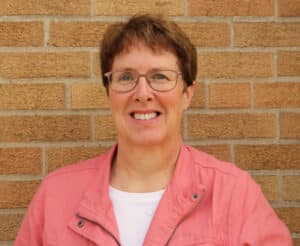 Denise Fase, a bridgebuilder of leaders, joins DVULI as part-time staff liaison, smiling in front of brick wall.